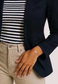 Silverarmband med en cirkulär berlock, bärs på en solbrun hand. Bakgrunden visar en marinblå jacka och en randig skjorta. En synlig knapp på byxorna.