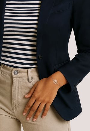 Braccialetto d'argento con un ciondolo circolare, indossato su una mano abbronzata. Lo sfondo mostra una giacca blu scuro e una camicia a righe. Bottone visibile sui pantaloni.