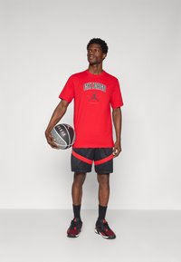 Červené bavlněné tričko s grafikou "CHICAGO", černé síťované šortky s červenými akcenty, černé ponožky a červené a černé sportovní boty. Drží basketbal.