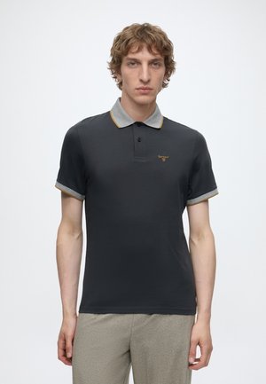 Jeune homme aux cheveux bouclés et bruns, portant un polo noir avec un col et des manches gris, debout sur un fond blanc uni.