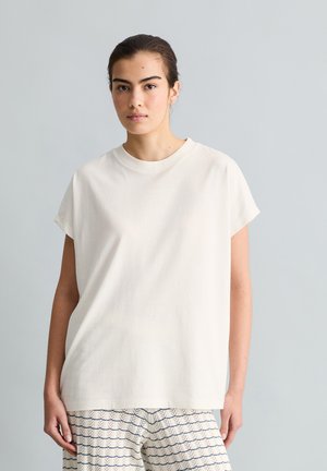 Junge Frau mit dunklen Haaren in einem weißen kurzärmligen Oberteil und gemusterten Hosen vor einem einfarbigen hellgrauen Hintergrund.