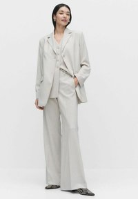 Vaaleanharmaa kolmoispuku, jossa on räätälöity bleiseri, yhteensopiva liivi ja levenevät housut. Kangas näyttää sileältä, suurilla nappeilla ja sivutaskuilla.