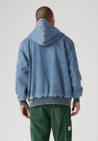 Haut à capuche en denim bleu clair avec une capuche structurée et des poignets côtelés. Présente un ourlet froncé et un tissu texturé. Porté avec un pantalon en velours côtelé vert.