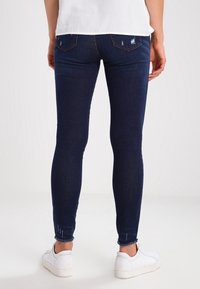 Jeans skinny azul oscuro con un diseño desgastado, con bolsillos traseros y dobladillos deshilachados, combinados con zapatillas blancas.