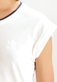 Bílý bavlněný tričko s námořnickým ribovým výstřihem, krátkými rukávy s ohrnutím a decentním bílým logem adidas potištěným na hrudi.