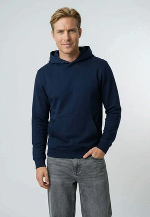 Blond man wearing a navy blue hoodie and gray jeans, standing with one hand in pocket, against a plain light background.