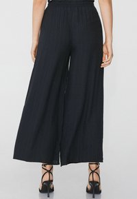 Pantalones negros de pierna ancha con una tela texturizada, que presentan una cintura elástica, un corte holgado y un elegante dobladillo abierto, combinados con tacones negros con tiras.