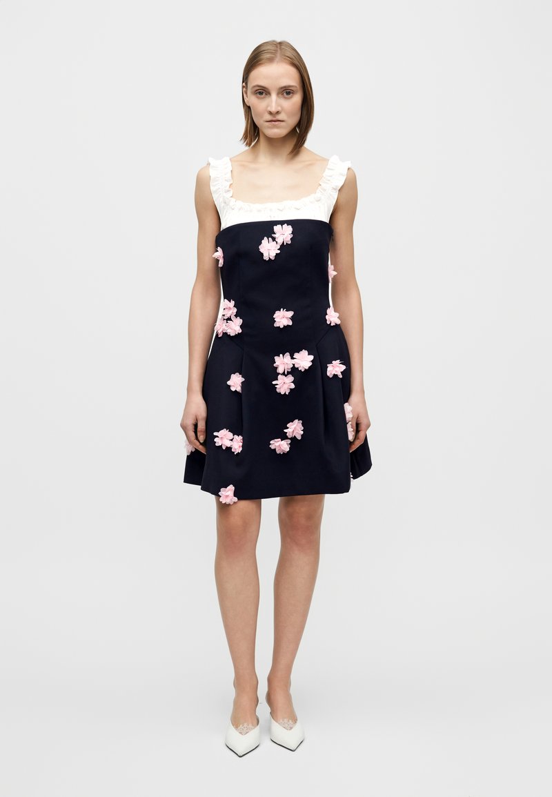 Vestito blu navy con scollatura bianca arricciata, decorata con applicazioni floreali rosa, con corpetto aderente e gonna svasata.