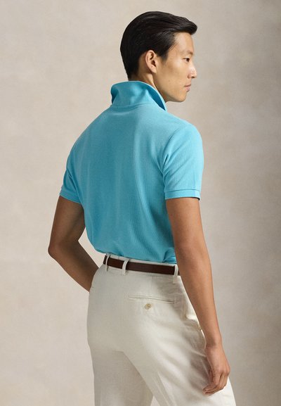 Ανοιχτό γαλάζιο πόλο με διπλωμένη γιακά, υφασμάτινη υφή, κοντά μανίκια και μπεζ παντελόνι με καφέ ζώνη. Πλάγια όψη από πίσω.