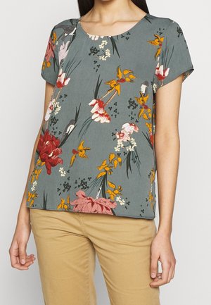Grünes Kurzarmtop mit rotem, gelbem und weißem Blumenmuster, getragen zu beigefarbenen Hosen von einer Person, die vor einem einfarbigen Hintergrund steht.