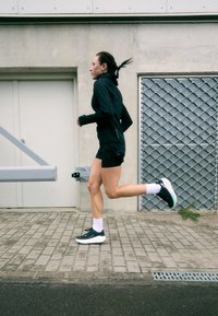 Čierna športová bunda a šortky, biele ponožky po kolená a čierne bežecké topánky. Beton pozadie s kovovou mriežkou. Osoba v pohybe.