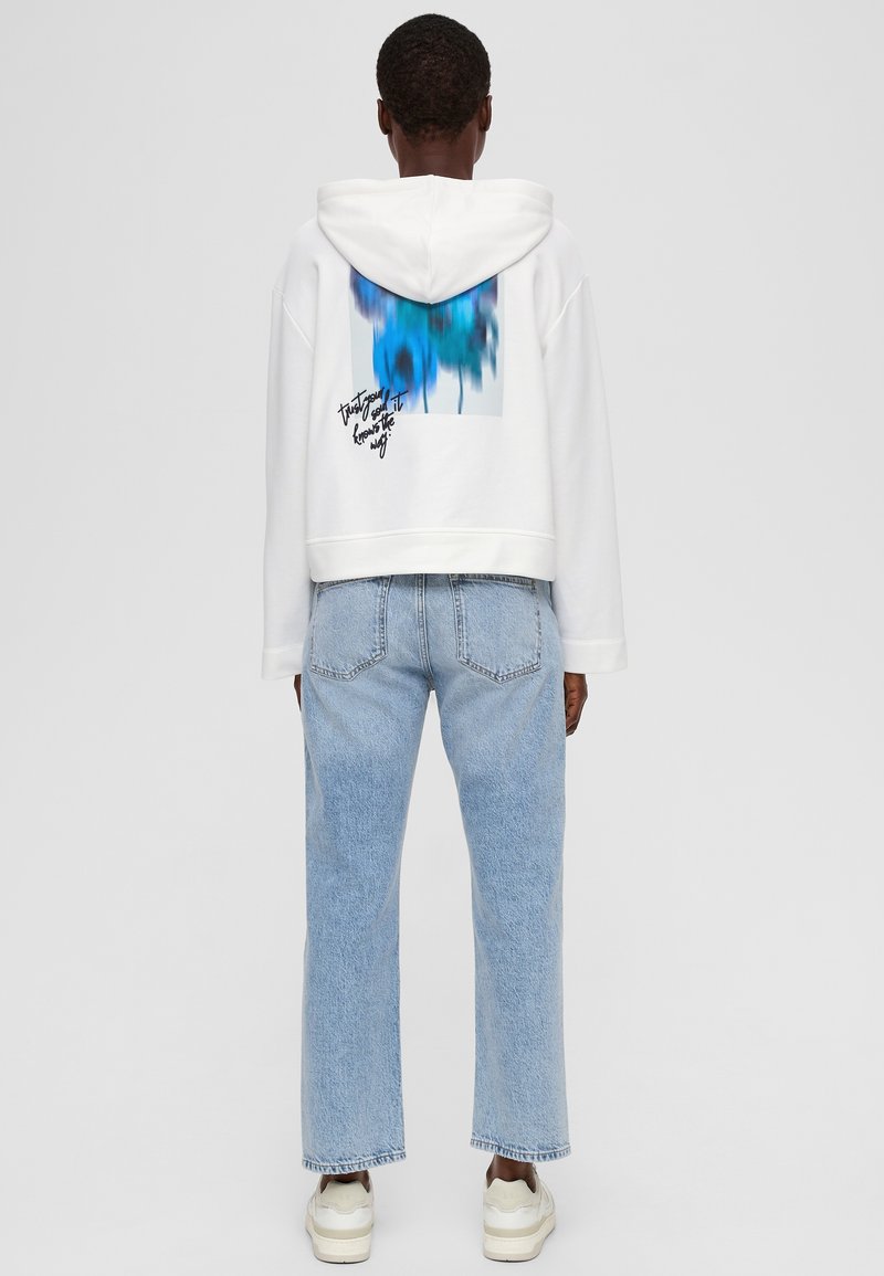 Weißer Cropped-Hoodie mit einem blauen abstrakten Design auf der Rückseite, kombiniert mit hellblauen Straight-Leg-Jeans und weißen Sneakern.