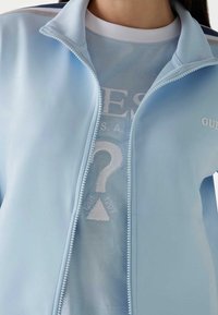 Ljusblå zipjacka i mjukt tyg med vit brodyr och en frågeteckensgrafik på det innersta lagret.