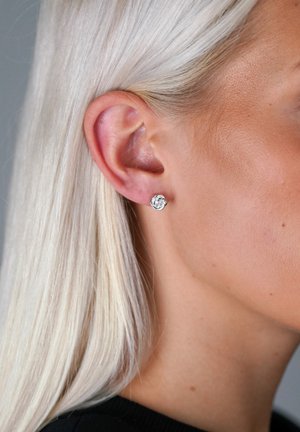 Silverringar i form av stift med en cirkulär design, med en klar ädelsten i mitten, placerade mot en ljus hudton och blont hår.