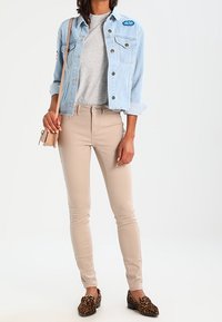 Giacca di jeans blu chiaro con dettagli in patch, T-shirt grigia, jeans skinny beige e mocassini maculati marroni, completando un outfit informale.