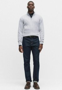 Jasnoszary sweter w splot kablowy z zamkiem przy kołnierzu, zestawiony z ciemnoniebieskimi dżinsami i brązowymi skórzanymi butami. Model stoi na tle jednolitego tła.