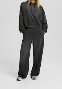 Felpa nera con cappuccio tagliata, con testo stampato "NYC"; abbinata a pantaloni da jogging neri larghi con cuciture laterali e tasche. Le scarpe includono sneakers grigie.