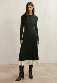 Robe en maille vert foncé à texture côtelée, manches longues et ourlet ivoire. Portée avec des bottes noires montantes et une silhouette ajustée.