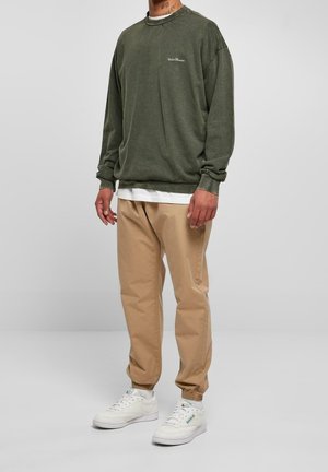 Mand iført olivengrøn sweatshirt, hvid undertrøje, beige joggerbukser og hvide sneakers, stående mod en ensfarvet grå baggrund.