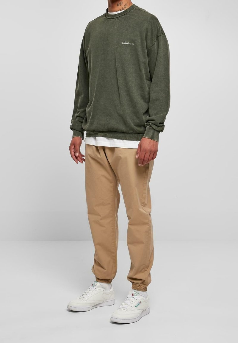 Hombre vestido con sudadera verde oliva, camiseta blanca debajo, pantalones joggers beige y zapatillas blancas, de pie contra un fondo gris liso.