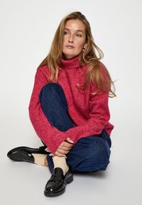 Sarkans augstais apkakles džemperis ar faktūru adījumu, apvienots ar brīviem zilajiem džinsiem un spīdīgām melnām mokasīnām. Silts krāsu palete un brīva fit.