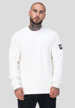 Hvid ribbet sweater med lange ærmer, rund halsudskæring og en sort lap på venstre arm. Bæres over mørke jeans.