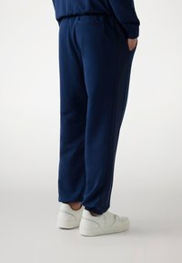 Marineblauwe sweatpants met een elastische tailleband, zijpanelen van gaas en opgebonden enkels, gecombineerd met witte sneakers op een lichte achtergrond.