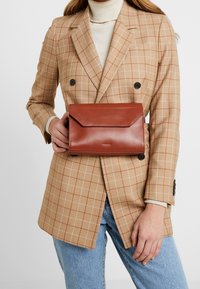 Clutch in pelle marrone con chiusura a pattina, texture liscia, forma rettangolare e logo impresso, appoggiato su un blazer a quadri beige.
