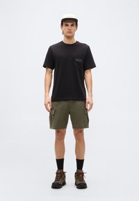 Νεαρός άνδρας που φοράει μαύρο μπλουζάκι Patagonia, χακί cargo σόρτς, μποτάκια πεζοπορίας, μαύρες κάλτσες και μπεζ καπέλο, στέκεται μπροστά σε λευκό φόντο.