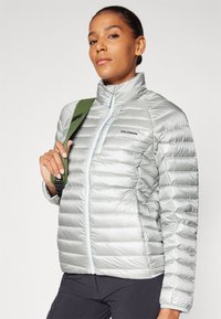 Ezüst színű pufferek jacket magas gallérral, steppelt kialakítással és világoskék cipzárral. A modell egy zöld pántot tart a vállán.