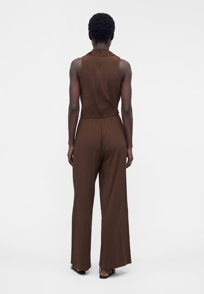Femme de dos portant une combinaison marron sans manches avec jambes larges et des sandales noires sur un fond gris uni.