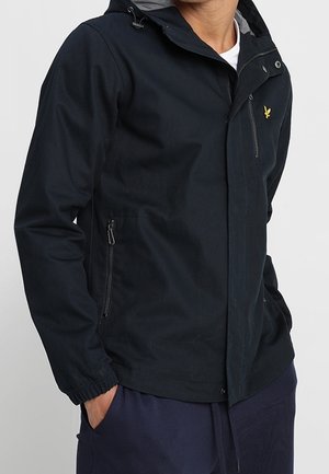 Mand iført en marineblå hættetrøje med lynlås og en gul fuglelogo på brystet, hænderne i lommerne, foran en ensfarvet baggrund.