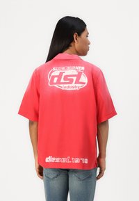 Camisa de manga corta, de corte holgado, en tejido rojo brillante con texto gráfico blanco en la parte posterior y el bajo. Cuello plano y aberturas laterales.