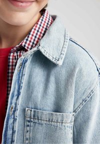 Jasnoniebieska kurtka jeansowa z klasycznym kołnierzem, z kieszenią na piersi i widocznym przeszyciem, noszona na czerwonej koszuli w kratę.