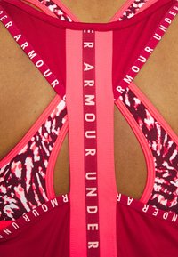 Gros plan d'une personne portant un soutien-gorge de sport rouge et rose Under Armour avec des bretelles à motifs et un ruban vertical marqué d'une marque dans le dos.