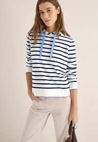 Femme portant un sweat à capuche blanc avec des rayures horizontales bleu marine et des cordons bleus, pantalon beige, tenant un sac à main marron sur un fond neutre.