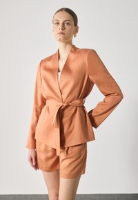 Orange satin blazer with a tied waist, featuring long sleeves and a deep V-neck, paired with matching shorts in a similar fabric.