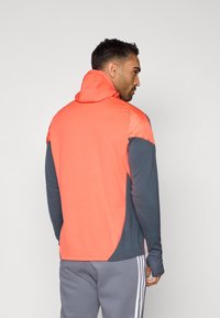 Φούτερ με κουκούλα που διαθέτει κοραλί πάνω μέρος και σκούρους γκρίζους μανίκους, κατασκευασμένο από ελαστικό ύφασμα. Περιλαμβάνει τσέπη καγκουρό και υφασμάτινες λεπτομέρειες.