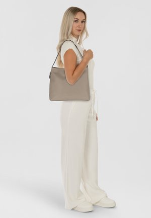 Beige Lederhandtasche mit glatter Textur und einem einzelnen Schulterriemen, getragen von einem Modell in einem weißen Outfit. Schlichtes Design, keine sichtbaren Beschläge.