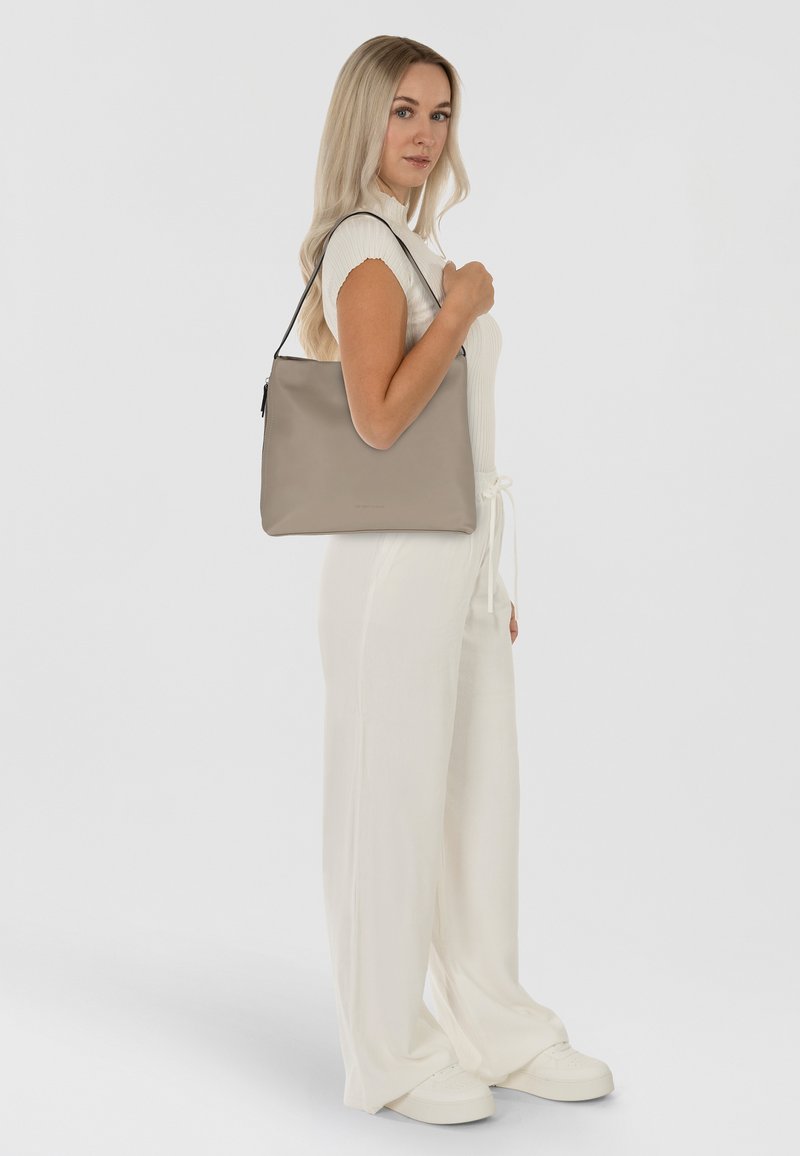 Beige leren handtas met een gladde textuur en een enkele schouderriem, gedragen door een model in een witte outfit. Eenvoudig ontwerp, geen zichtbare hardware.