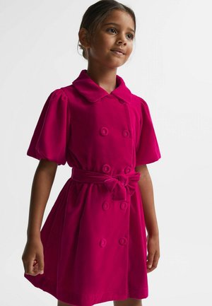 Jong meisje draagt een felroze dubbelgeknoopt jurk met pofmouwen en een gestrikte riem, staand tegen een effen lichte achtergrond.
