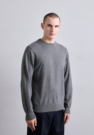 Un jeune homme aux cheveux courts portant un pull gris uni à col rond et un pantalon sombre, se tenant contre un mur texturé de couleur gris clair.