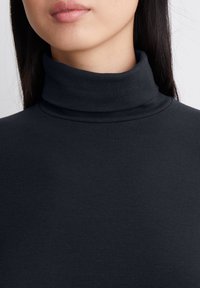 Donna che indossa un maglione nero a coste con colletto alto, che mostra il collo, il mento e le labbra su uno sfondo bianco.