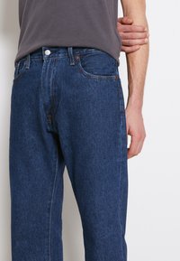 Dunkelblaue Denimjeans mit geradem Schnitt, ausgestattet mit kupferfarbenen Nieten und einem klassischen Knopfverschluss. Tragen mit einem grauen T-Shirt.