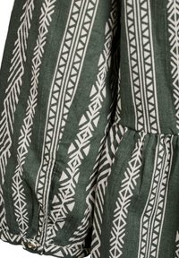 Grön duk med vita geometriska mönster, med en samlad söm och knappdetalj vid fållen. Bomullsmaterial med en mjuk textur.
