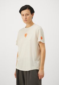 Svjetlosmeđi pamučni majica kratkih rukava, s izvezenim narančastim logom lava na prsima i crvenim Nike swooshom na rukavu.