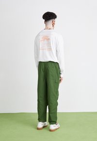 Vit långärmad skjorta med en rosa utomhusgrafik på baksidan, ihop med gröna cargobyxor och vita sneakers.