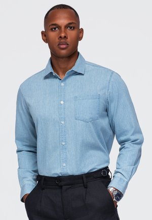 Homme les mains dans les poches, portant une chemise bleu clair boutonnée et un pantalon noir, debout devant un fond clair uni.