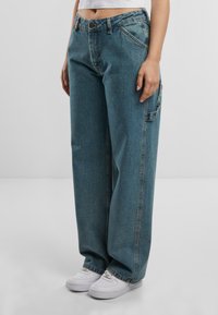 Blugi din denim albastru, cu croială lejeră, talie înaltă și buzunar lateral. Prezintă cusături clasice și textură uzată. Asortați cu adidași albi.