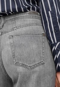 Grijze spijkerbroek met een achterzak, doorgestikte details en een merklabel. Draagt bij een gestreept marineblauw shirt.